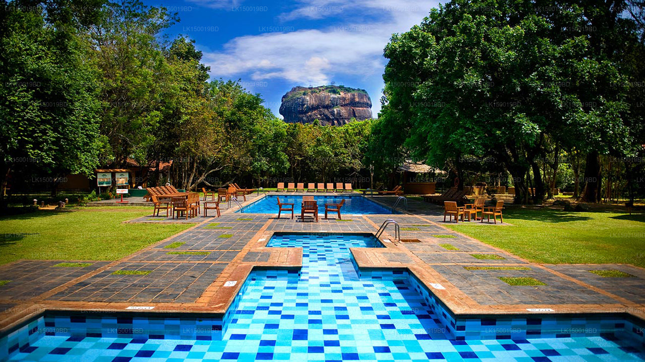 The image depicts the Sigiriya Village Hotel, which is set in a scenic location with a pool and cottages overlooking the Sigiriya Rock Fortress. The background includes lush greenery and a clear blue sky.